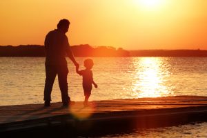Silhouette of father and son walking on pier holding hands with sun in background